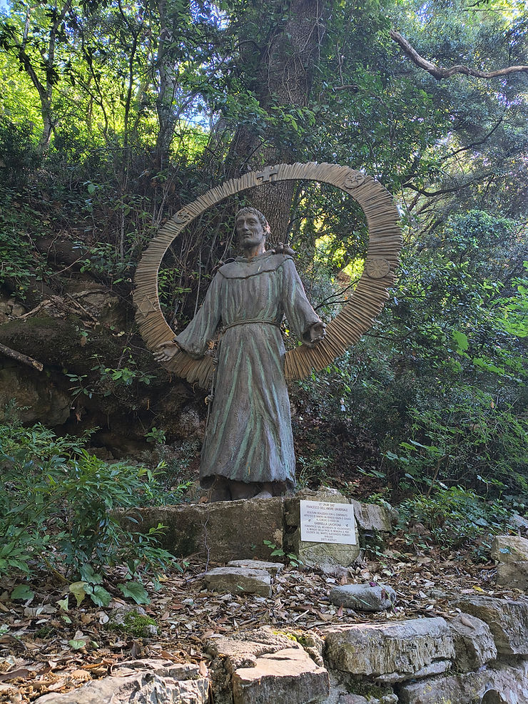 Stone saint statue in a leafy forest, framed by a circular halo, with an Italian plaque at the base.